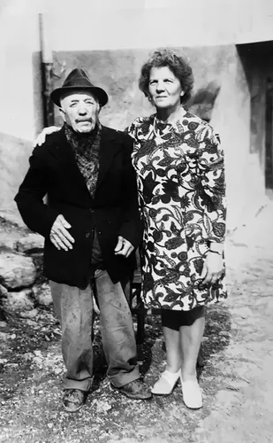 Photo de famille : un vieil homme au chapeau et une femme en robe fleurie devant une maison valdôtaine